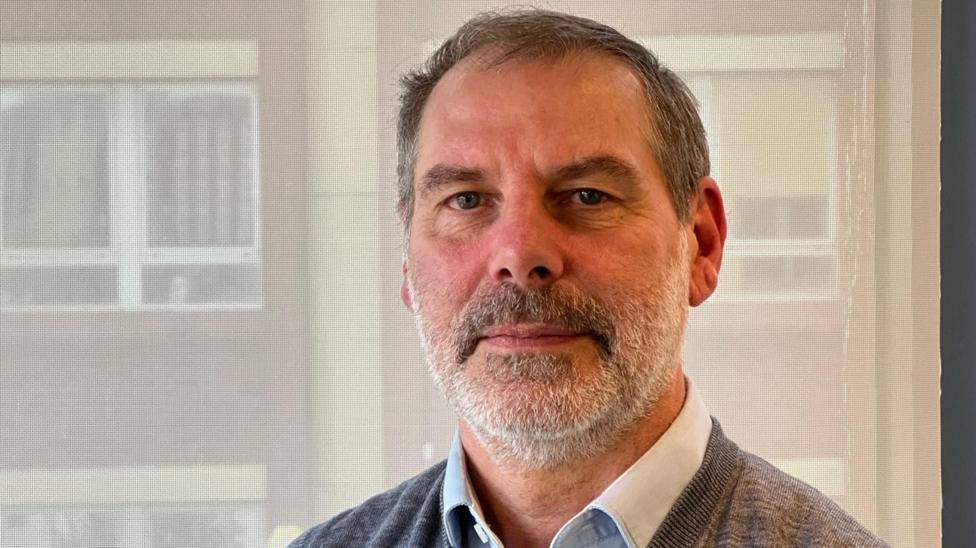 Head-and-shoulders portrait of Darren Squires, from the Hull Food Partnership. He has short, dark hair and a grey beard, wearing a light blue collared shirt under a grey sleeveless sweater. He is facing the camera indoors, standing in front of large windows with a softly blurred office-style background and natural light illuminating the scene.