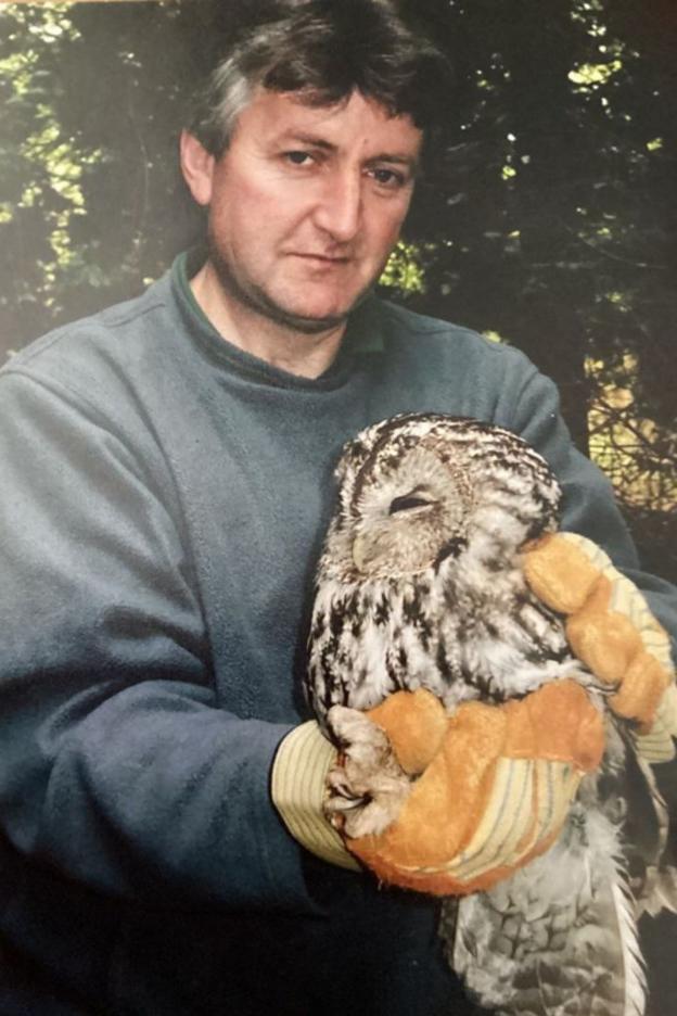 A photograph of a man holding an owl. He is wearing a green jumper with brown gloves on. The photograph was taken 20 years ago.