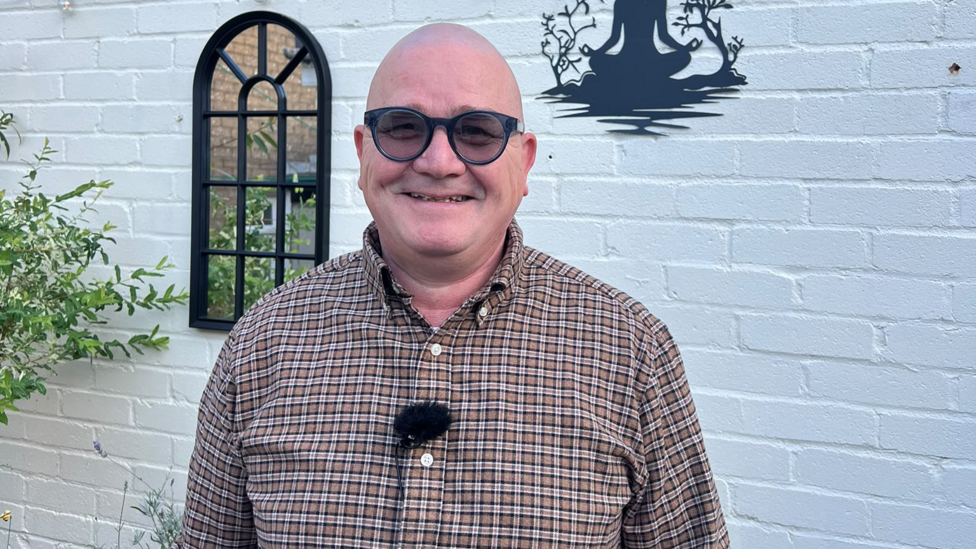 Paul Scanlon - a middle-aged bald man wearing a long sleeved checked shirt with a small microphone clipped to one of his buttons. Paul is wearing dark, circular glasses that are slightly tinted and he is standing in front of a whitewashed brick wall. 