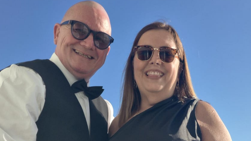 Paul Scanlon and his wife Deborah standing outside under a clear blue sky. Paul is wearing dark, circular glasses, a white shirt, waistcoat and a black bow tie. Deborah has horn-rimmed Ray-Ban sunglasses on and has dark shoulder-length hair. She is wearing a dark, over-the-shoulder dress and is slightly poking her tongue out between her teeth