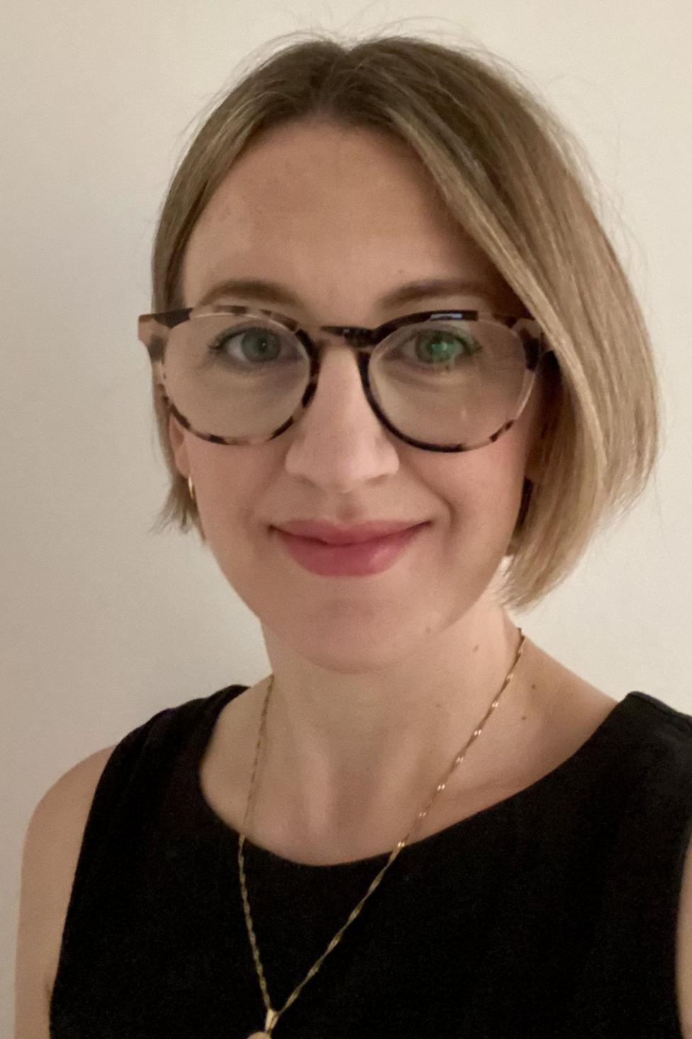 Lisa Barker is a woman wearing a black round-necked top with no sleeves. She is wearing a gold necklace and tortoiseshell glasses and has light brown bobbed hair.