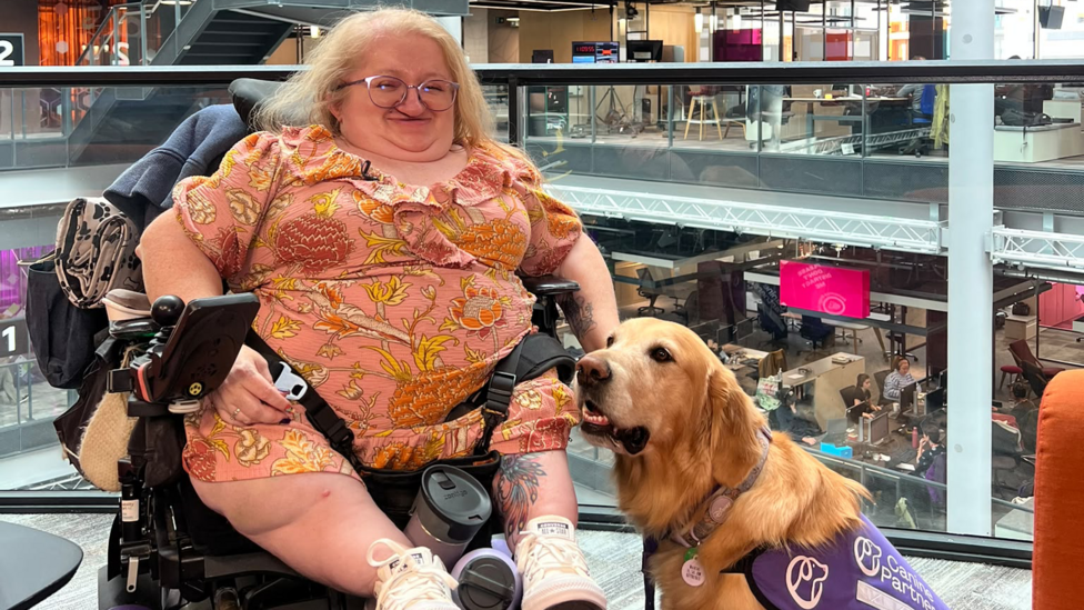 Kat Watkins is sitting in her wheelchair inside an office building with a golden retriever dog next to her wearing a purple vest that says canine partner. Kat has shoulder length blonde hair and is wearing glasses and a peach coloured dress covered in a floral pattern. She has a tattoo on the inside of her left leg and has on all white Converse trainers.