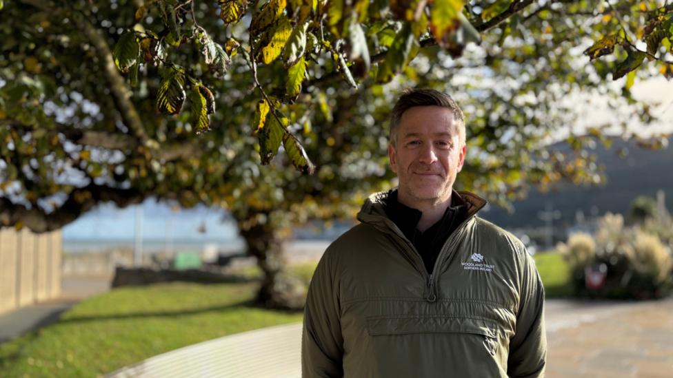 John Martin standing in front of the tree. He has short brown hair, wearing a khaki green jacket.