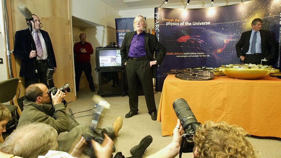 Pillinger is stood in front of gather photographers while stood next to a model of Beagle 2