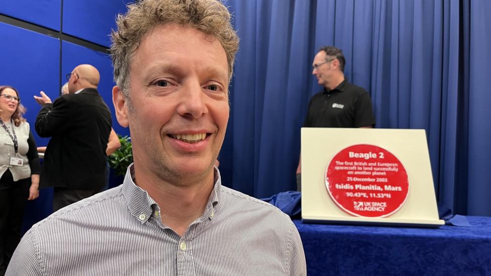 A man is smiling with a red plaque on display behind him detailing Beagle 2