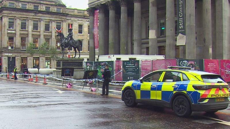 A police cordon surrounds a statue of the Duke of Wellington on horseback. The statue has three orange cones on it. A police car and a police officer on foot are on a wet street outside a columned building