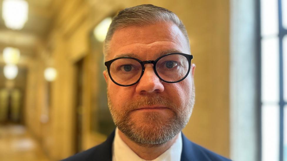 Colin McGrath has short dark hair and beard and is wearing black glasses.