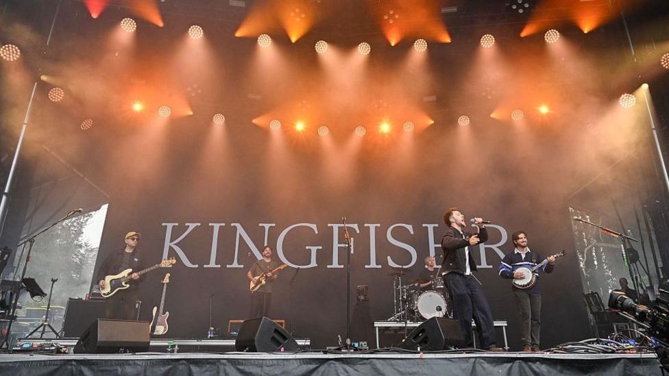 A wide shot of five men with guitars, performing on a lit up outdoor stage. The logo KINGFISHR is projected behind them.