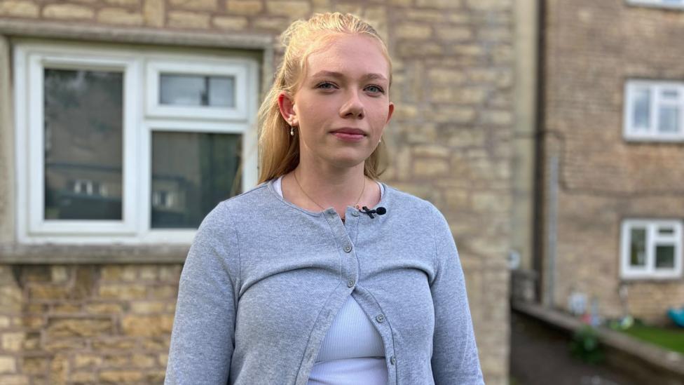 A woman with long straight blonde hair looks into the camera with a slight smile. She wears a light blue vest top with a great cardigan over the top. She stands in front of a house.