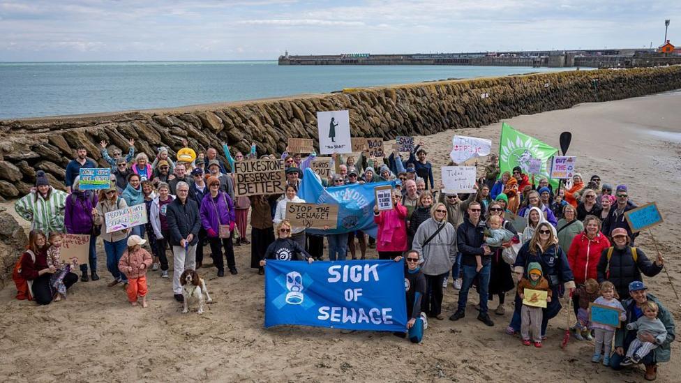 A group of people stood on a sandy beach. They are holding signs and banners voicing their displeasure against the sewage in UK waters.