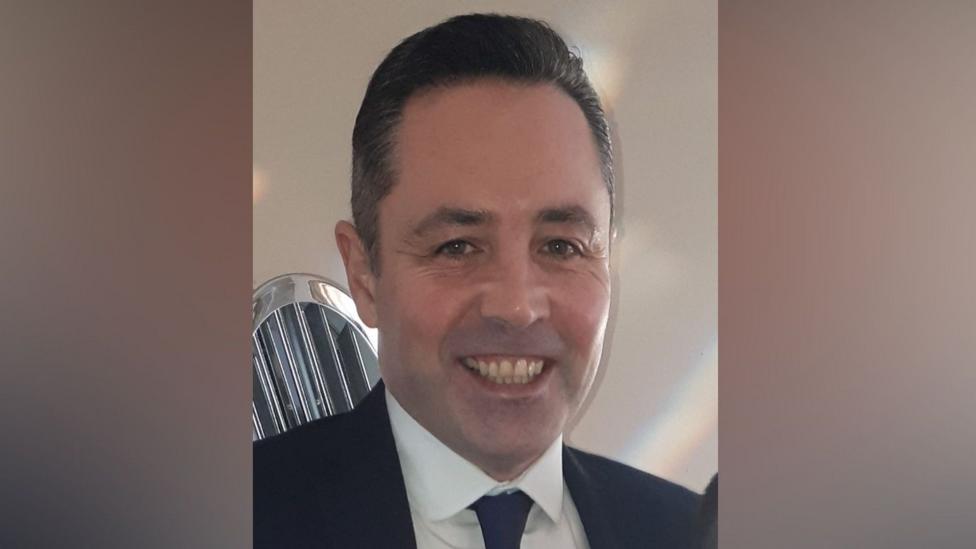 Eamonn Moore has short black hair and is wearing a black suit