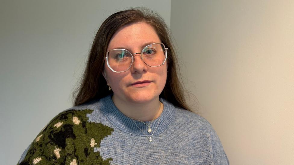 Jennifer, a woman with long brown hair and glasses looking at the camera. She wears a blue jumper with a green shape on the shoulder.