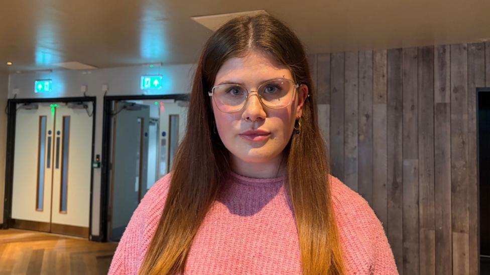 Megan, who is wearing a pink jumper and glasses. She has long dark hair and has an eyebrow and nose piercing.