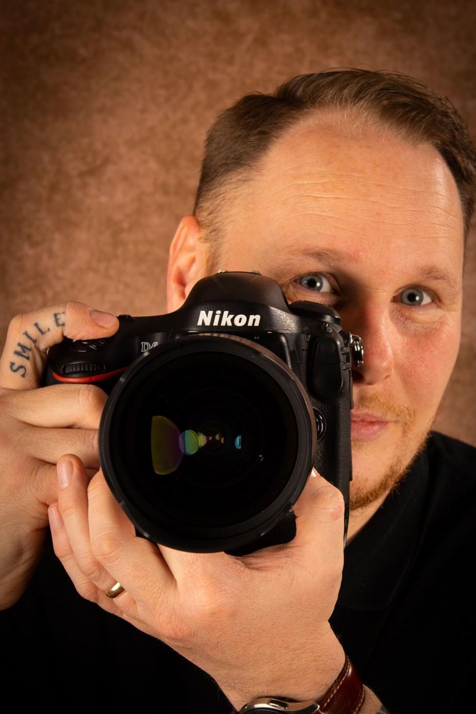 Ian holding a Nikon black camera. You can see him behind the camera. He has short brown hair and blue eyes.