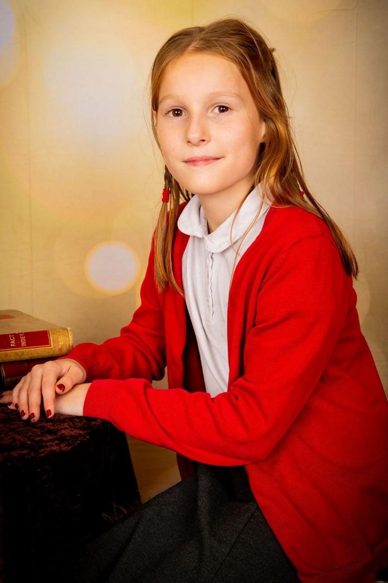 A girl posing for a school photo. She has shoulder-length brown hair and a red cardigan, white t-shirt and black trousers. Her hands are on a table.