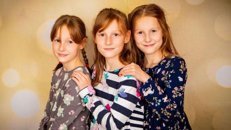 Three girls posing for a photo. They are have auburn hair and smile at the camera. The one on the right has a blue dress with flowers, in the middle she wears a black and white striped top with flowers and the girl on the left has a brown dress with flowers