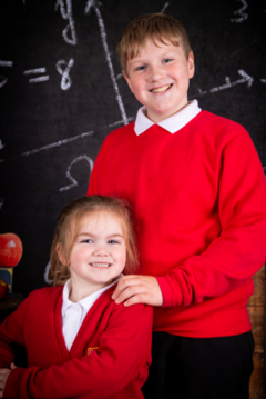 A boy and a girl posing for a school photograph. They both have red jumpers/cardigans on and smile.