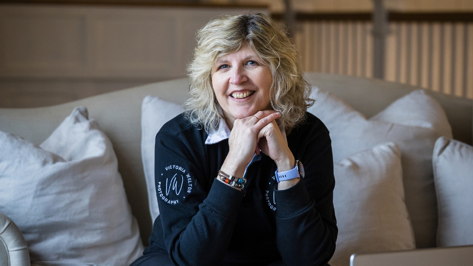 Victoria Welton is sitting on a sofa wearing a navy blue sweatshirt over a lighter-coloured shirt. She is a middle-aged woman with shoulder-length wavy blonde hair and she is smiling widely while looking directly at the camera. She is sitting on a cream-coloured sofa which is covered in large cream-coloured cushions.