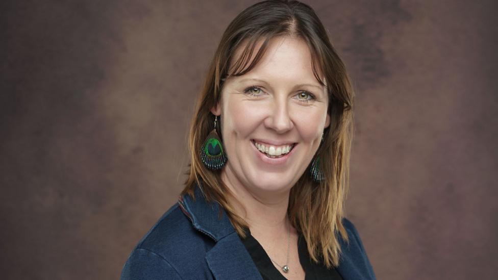 Tessa Chapman is seen from her shoulders up, smiling at the camera. She is wearing a blue blazer and feather earrings.