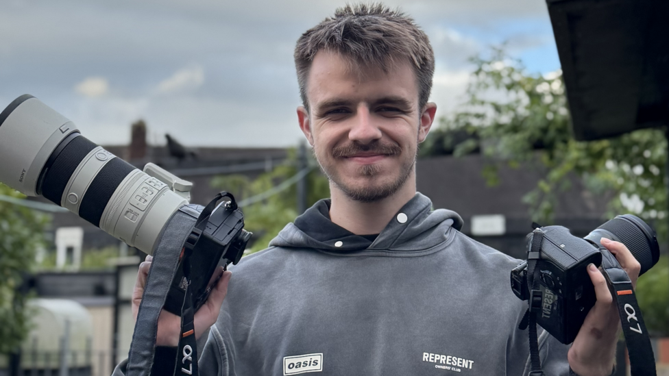 A man with short brown hair looks at the camera. He is holding two sports cameras, one in each hand and has a grey hoody on.