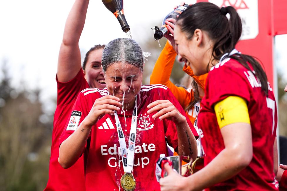 Footballers in a red football strip celebrating. They have medals on and one of the women is being doused in alcohol.