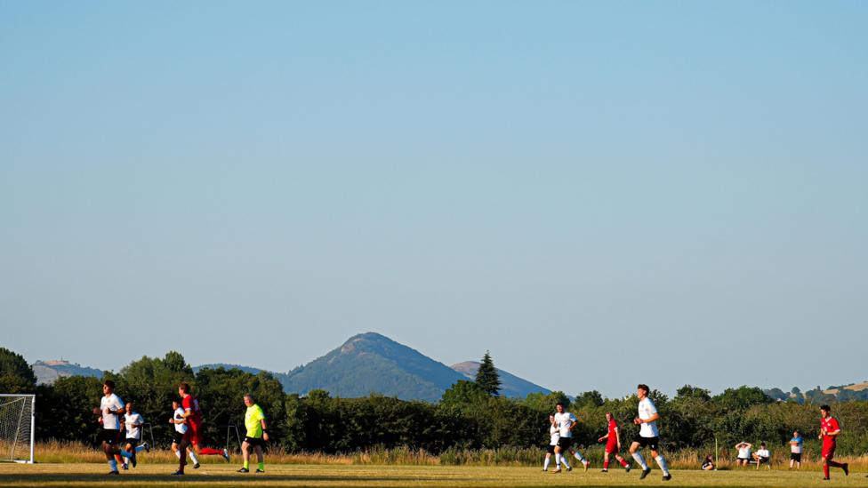 Footballers on a pitch in a white strip and a red strip along with a referee in fluorescent yellow and black kit. The background has mountains and trees. 