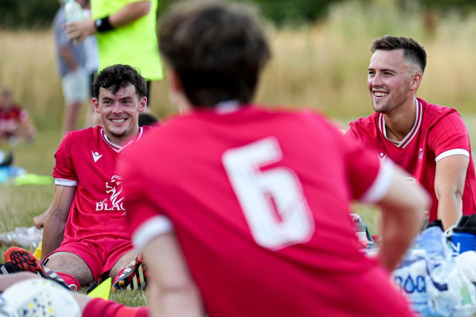 Footballers sat on the floor smiling. One has his back to the camera. While the other two are looking at the camera. The one with his back is slightly off focus while the others are in focus.