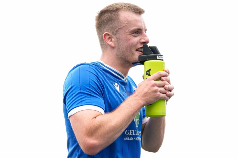 A man with a blue football shirt and short light-blonde hair drinking from a water bottle.