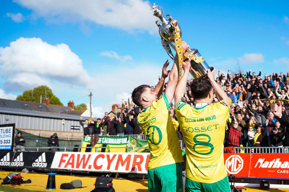 Two footballers in a yellow and green strip hold up a football trophy in front of a crowd.