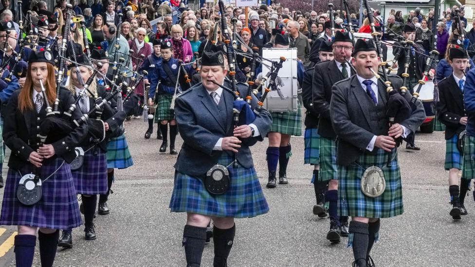 A piping band in green and purple kilts leads the procession of singers along a road in Fort William