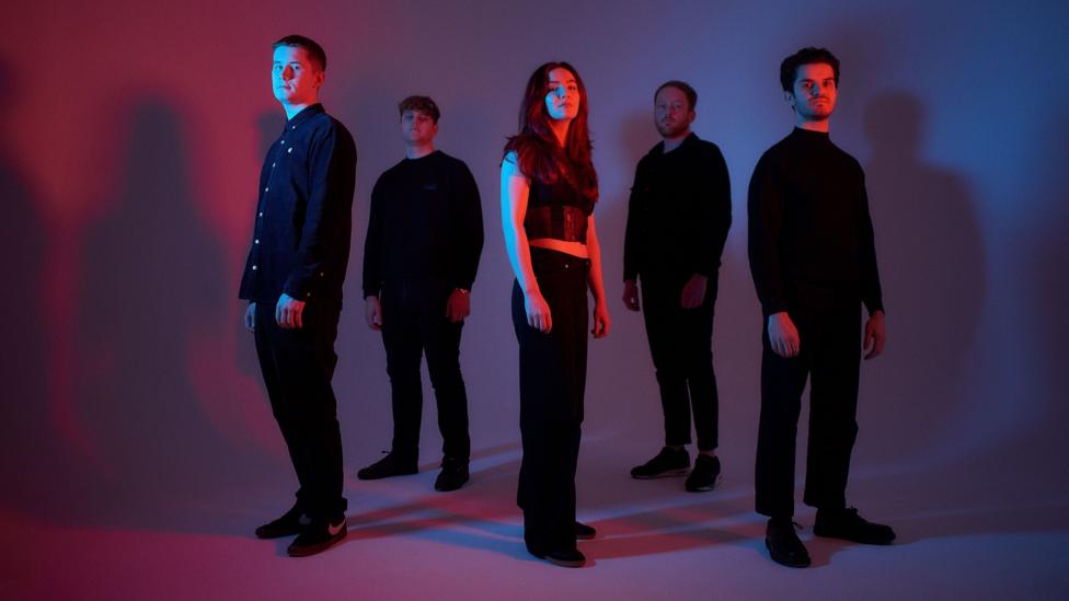 In a promotional photograph, the five members of Dlù stand in a red and blue lit room wearing black and holding a commanding pose and stare at the camera.