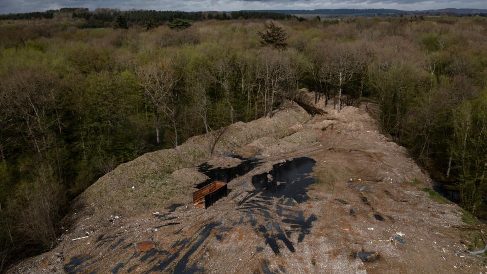 An aerial view shows a woodland clearing that is covered by dumped waste. The rubbish is piled high on the edge of the surrounding treeline and amid the waste are pools of dark water and an abandoned container. The trees stretch to the hills on the horizon, underneath a cloudy sky.