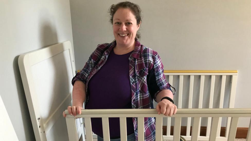 Jacqui Hunter, who is heavily pregnant. She is wearing a checked shirt and smiling at the camera and holding the sides of a baby cot that is being assembled