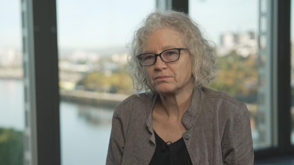 Professor Christine McCourt is sitting with a window behind her and an out-of-focus view of the River Clyde. She is wearing a black top and a beige jacket and black rimmed glasses.