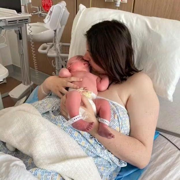 Claire Fleming in a hospital bed holding newborn baby Andrew who is wearing a nappy and hospital bands on his ankles.