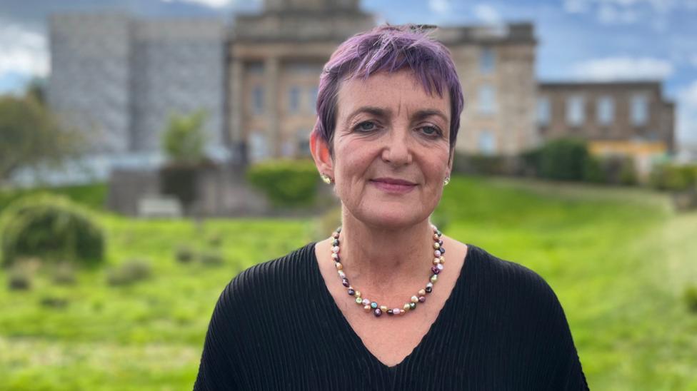 Health Secretary Angela Constance in a black top with a coloured necklace standing in a grassy area with a building out of focus in the background.