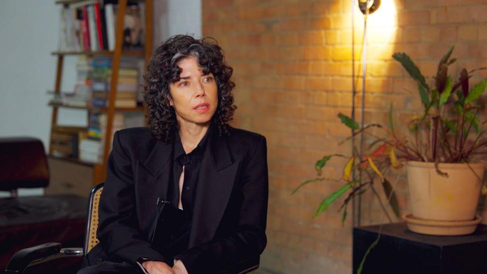 Meredith Whittaker seated, being interviewed. She is wearing a black top and black jacket and has dark, curly hair. She is indoors. Behind her is a bare brick wall and a bookcase. There is a pot plant on the right of the picture.