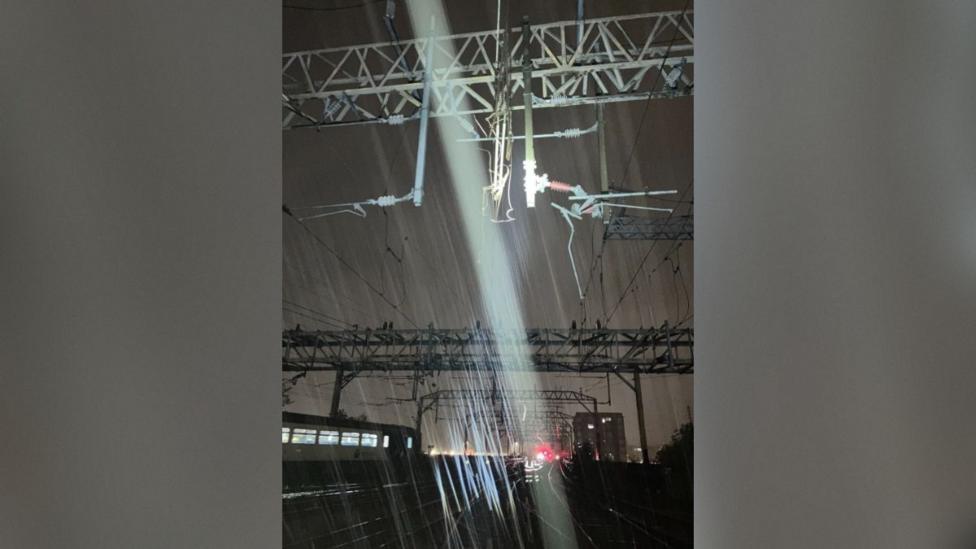 A night-time view along railway tracks beneath a series of large metal gantries supporting overhead electrical wires. Several cables and pieces of electrical equipment appear to be hanging from the structure above the tracks. A strong white light shines down through heavy rain.