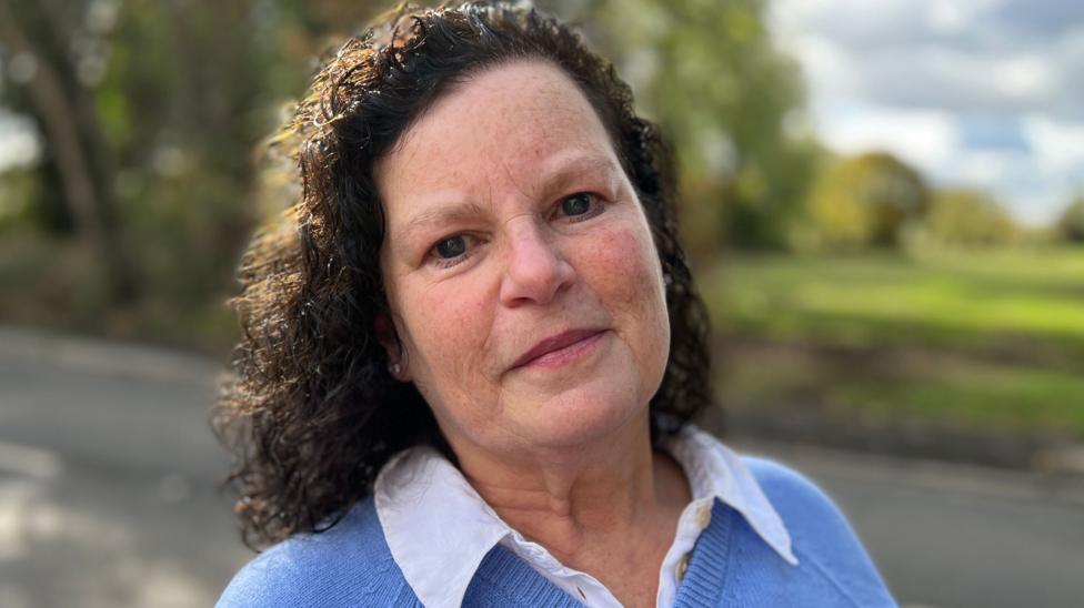 A woman with dark brown curly hair is looking straight at the camera. She is wearing a blue top with a white collar.