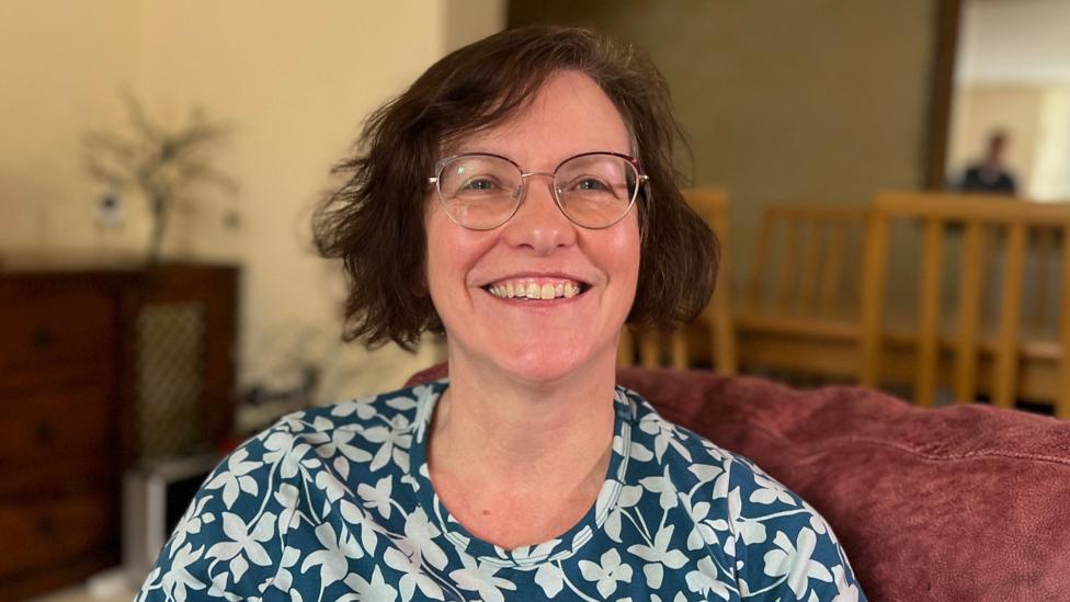 A woman with short brown hair and glasses is smiling at the camera. She is wearing a blue and white floral top.