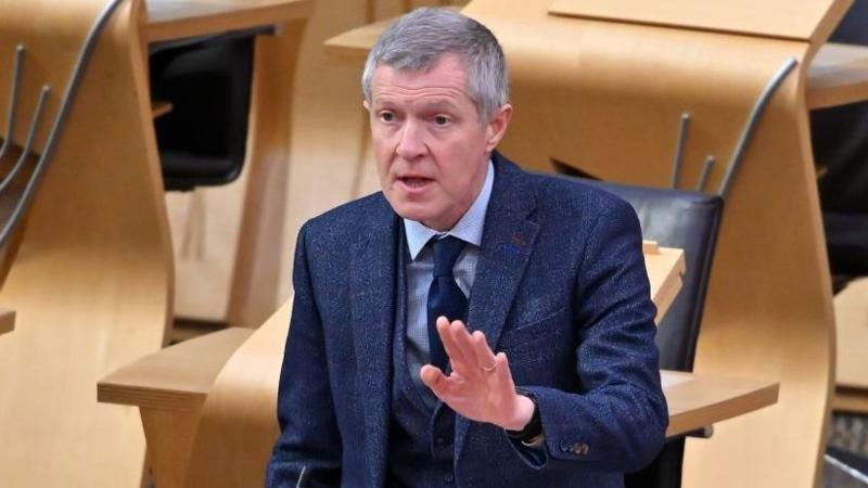 Willie Rennie in the Scottish Parliament chamber. He is standing and has his right arm raised slightly. He is wearing a blue chekced suit over a light blue shirt.