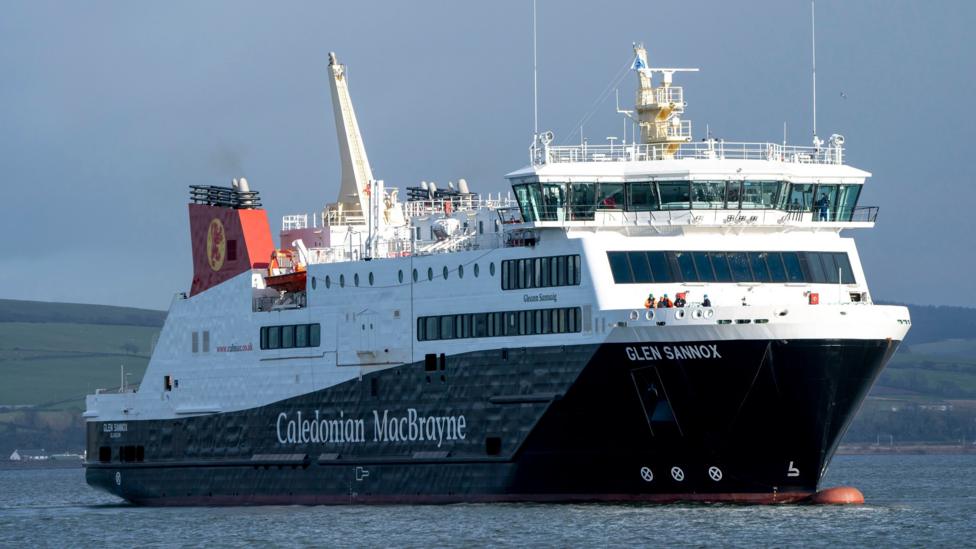 MV Glen Sannox on the water. It has a dark hull with the words "Caledonian MacBrayne" and "Glen Sannox" in white lettering. The top of the ship is white. The funnel is red.