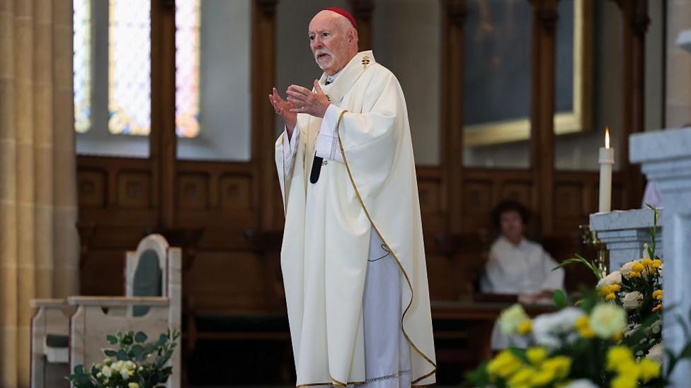 Archbishop William Nolan, standing and speaking while in church. He is wearing white robes, and has a white beard and hair.