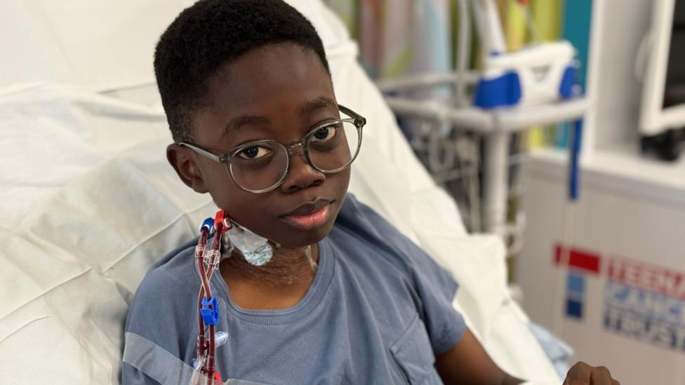 JoelSamuel sits up in bed at Birmingham Children's Hospital. Tubes collecting his cells are fixed to his neck. He is wearing glasses and a blue hospital gown.