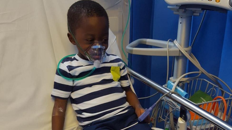 JoelSamuel when he was younger, sitting in a hospital bed being treated for the painful effects of sickle cell disease. He is wearing a navy and white striped T-shirt.