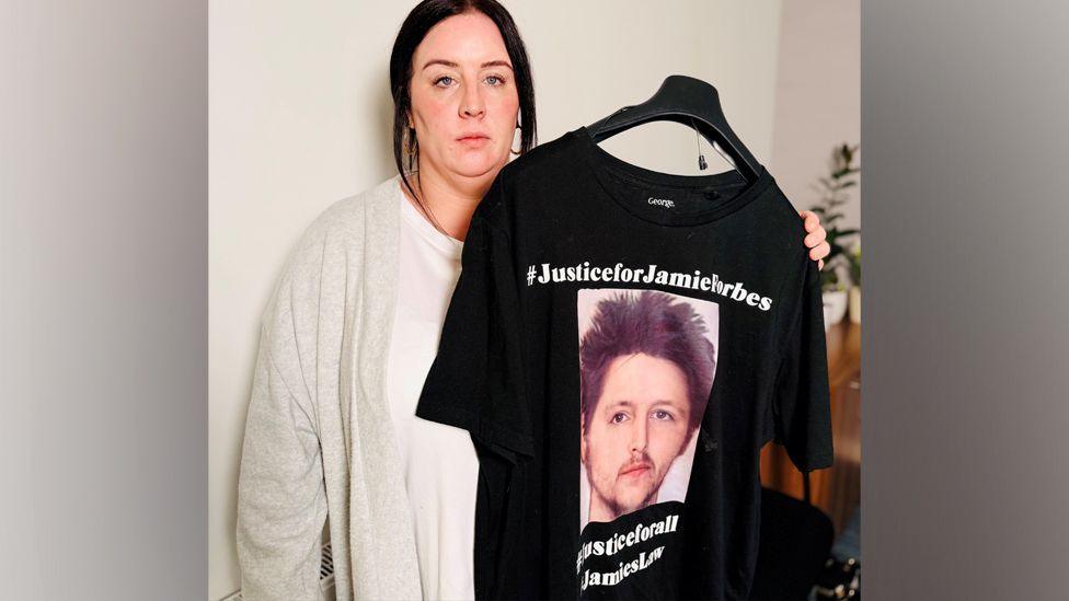 Lyndsay-Anne Forbes looking at camera and holding a T-shirt with a photo of her brother Jamie Forbes on it and words including #JusticeforJamieForbes