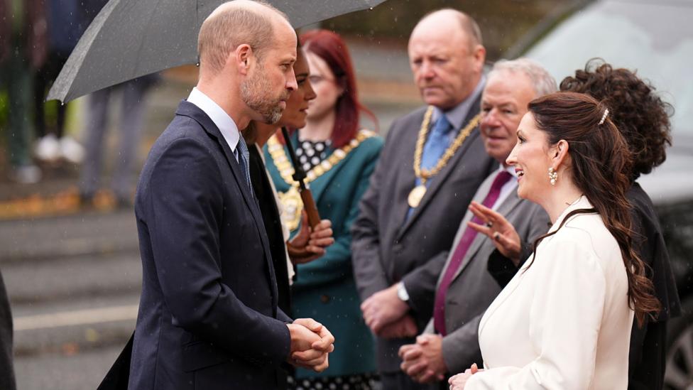 A man wearing a navy suit talks to a women wearing a white blazer, there are other people in the background. It is raining