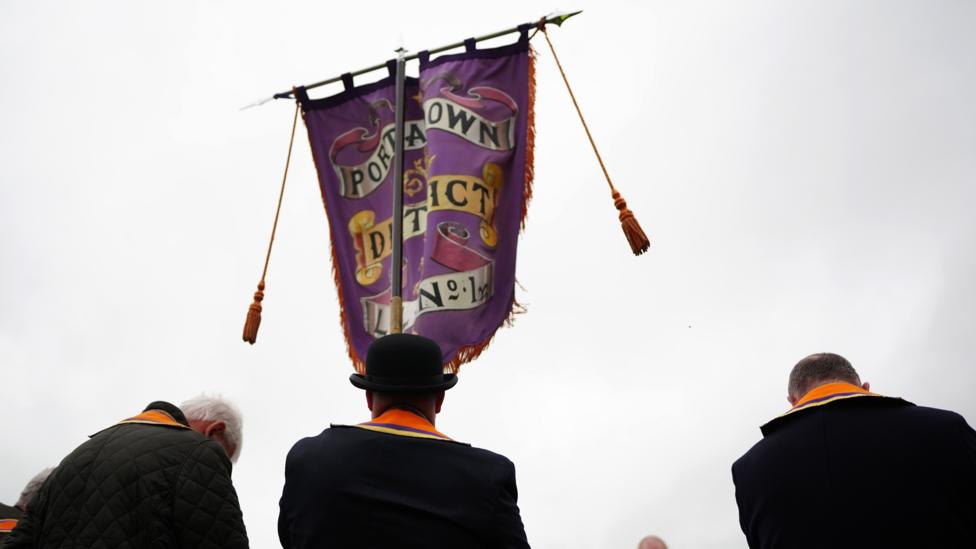 A group of Orangement in black suits and with Orange sashes standing with their backs to the camera, carrying a purple standard with 'Portadown District' emblazoned on it.