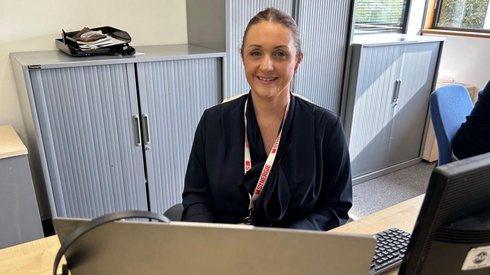 A woman sat behind a desk in an office. She smiles at the camera. She has a dark blue top on and a red and white lanyard.