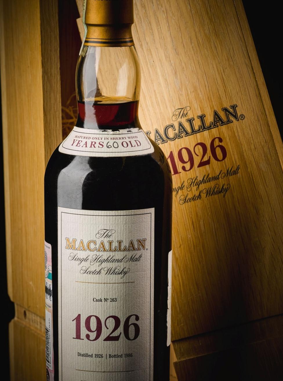 A bottle of Macallan 1926 whisky. It is a clear tall whisky bottle with a white label and stands in front of a wooden whisky box